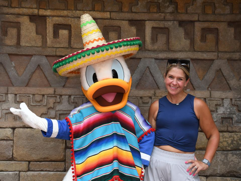 Terri Peters posing with someone dressed in a Donald Duck costume featuring a brightly colored striped poncho and a sombrero with orange, red, yellow, and green piping. Terri, who has blond hair, poses with her left hand on her hip and the other on the mascot's back. She has pink painted fingernails and wears a sleeveless navy-blue top that's cropped slightly at her waist, gray athletic shorts, and sunglasses on her head. They're in front of a brick wall with geometric designs.