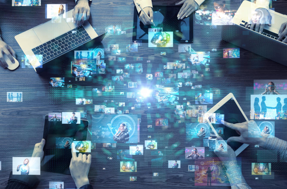 Employees sitting around table with computers out, and technology icons superimposed on table.