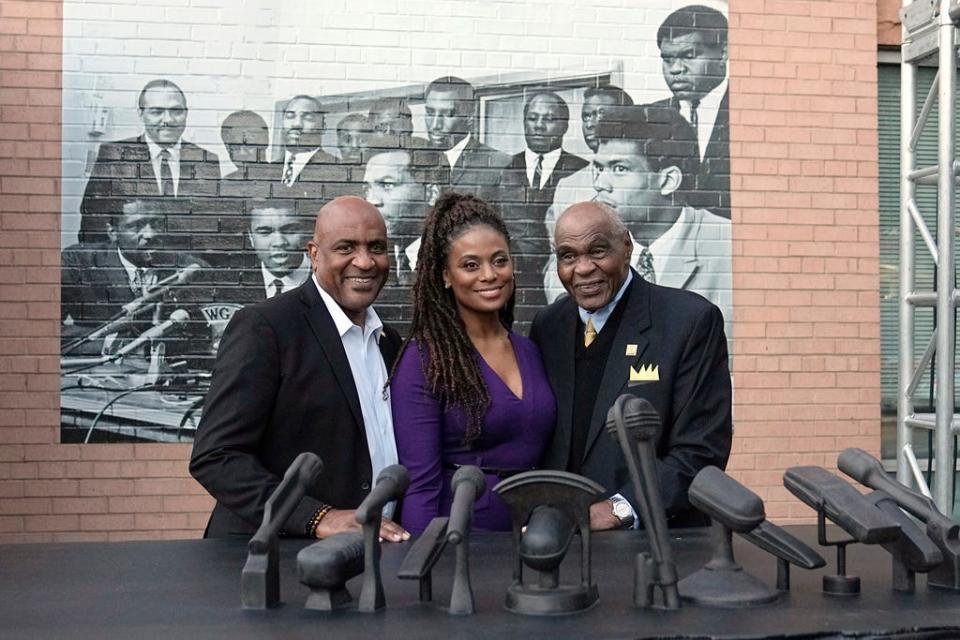 Kevin Clayton, left, Cleveland Cavaliers vice president of social impact and equity; Monique Brown, widow of Jim Brown; and John Wooten, right, pose for a photo behind a sculpture the 1967 Cleveland Summit, Wednesday, Oct. 11, 2023, in Cleveland. The sculpture is a representation of the press conference table where the Black athletes sat; a photo of the event is at rear. (AP Photo/Sue Ogrocki
