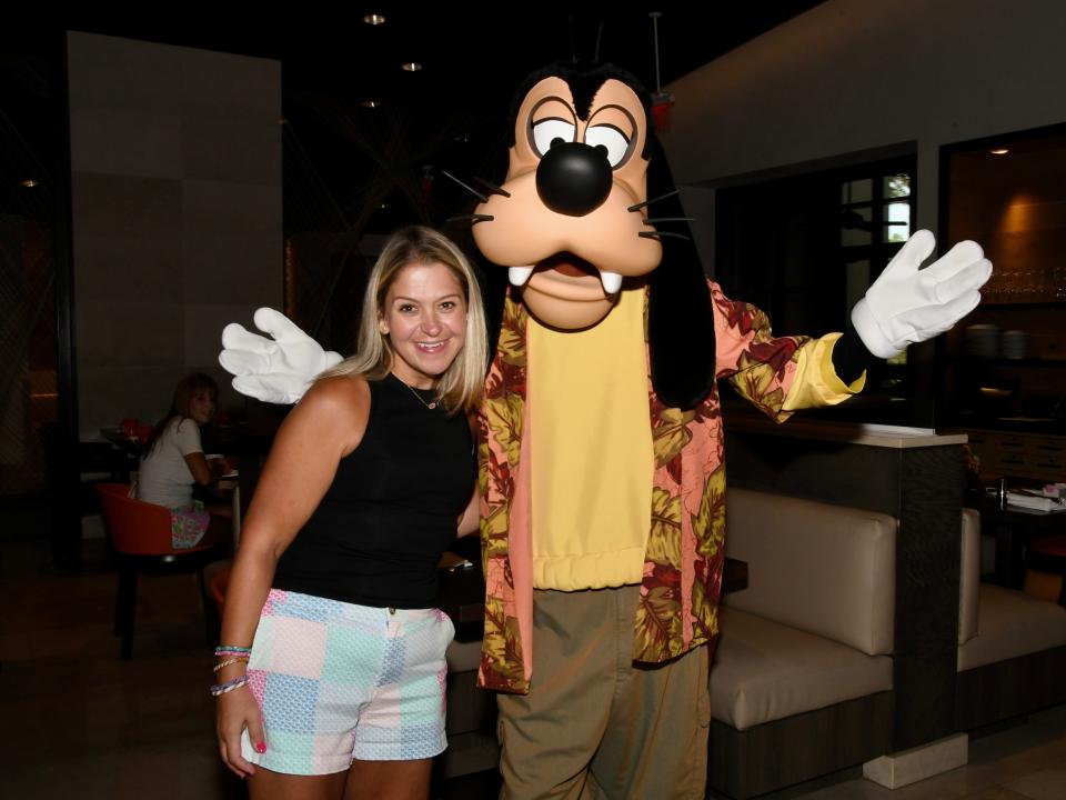 Terri posing with a Goofy mascot. She has blond hair and dark eyes and smiles widely. She wears a black sleeveless top and shorts with a blue, green, orange, and yellow patchwork print. Her left hand is around Goofy, who wears an orange Hawaiian shirt with yellow leaves on top of a yellow shirt, and green pants. His white-gloved hands are held out. Behind them is a booth at a restaurant.