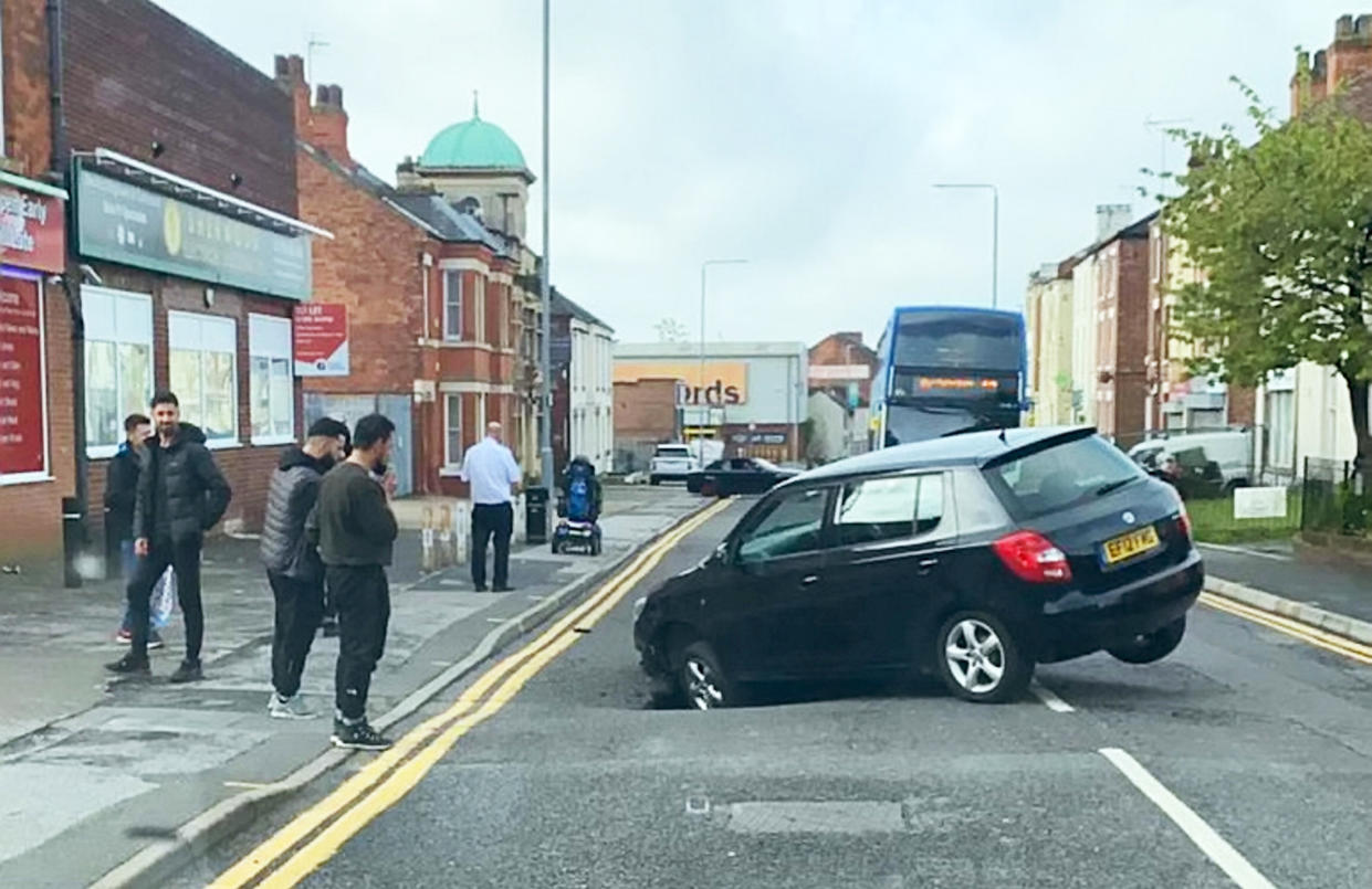 A massive sinkhole opened up in a busy street and swallowed a car causing cops to shut the road. See SWNS story SWLNsink.  The car was left wedged in the sinkhole with its front wheel trapped but incredibly no one was injured.  Road users on Carlton Road, Worksop, Notts., were forced to drive around the large sinkhole after it opened up at 9.00am on Saturday morning.  Cops were forced to close the road after the elderly Skoda Fabia driver accidentally trapped his car.  Locals said the area is prone to random holes forming as the road is built on top of an old drain.  Russell Gull, the owner of Res Barbers, helped the trapped driver out of his sunken car. 