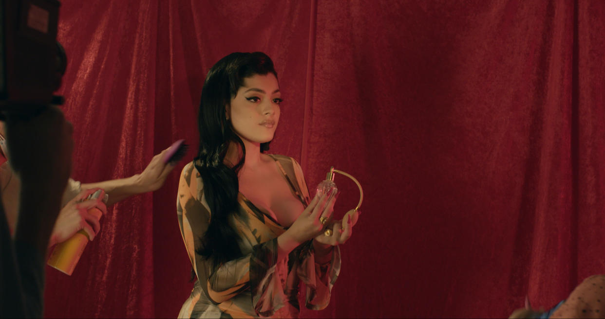 Jacks Haupt poses against a red velvet backdrop in a low-cut yellow dress with flowing sleeves.