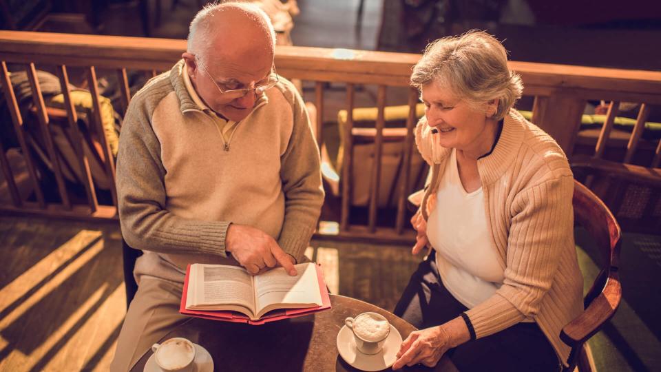 High angle view of a senior man reading a book to his wife in a cafe who is enjoying listening to him.