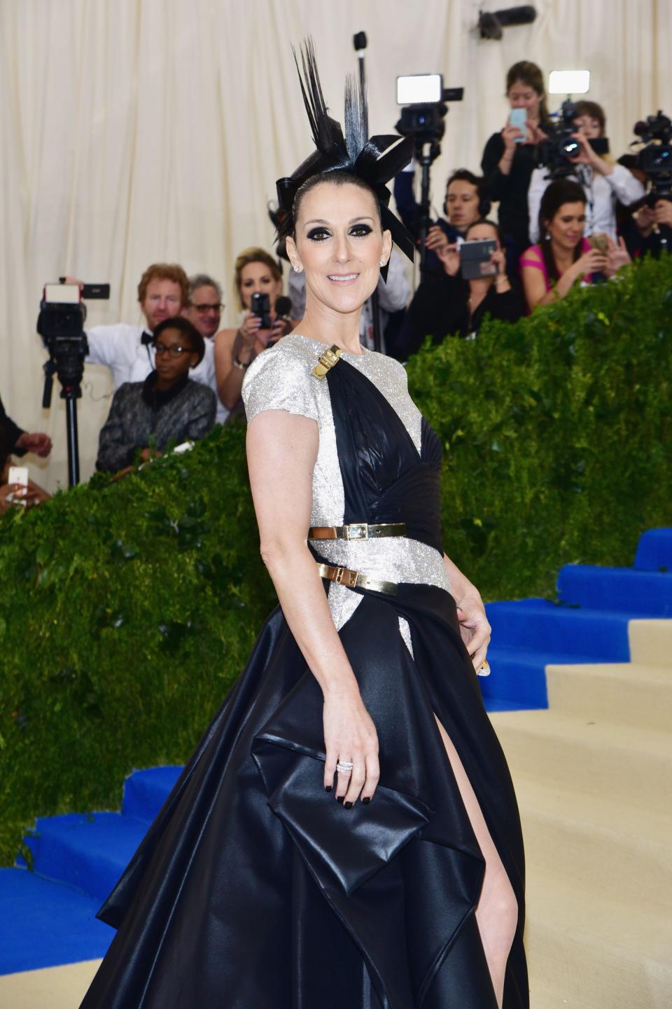 Celine in an asymmetrical shining black and silver short sleeve voluminous gown. She has dark eyeliner and is wearing a black pointed headpiece.