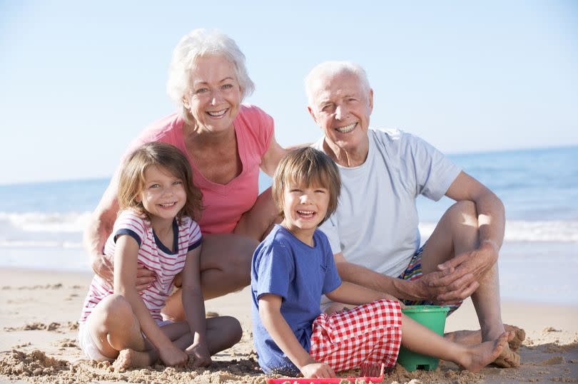 Grandparents And Grandchildren Building Sandcastle On Beach Smiling To Camera.