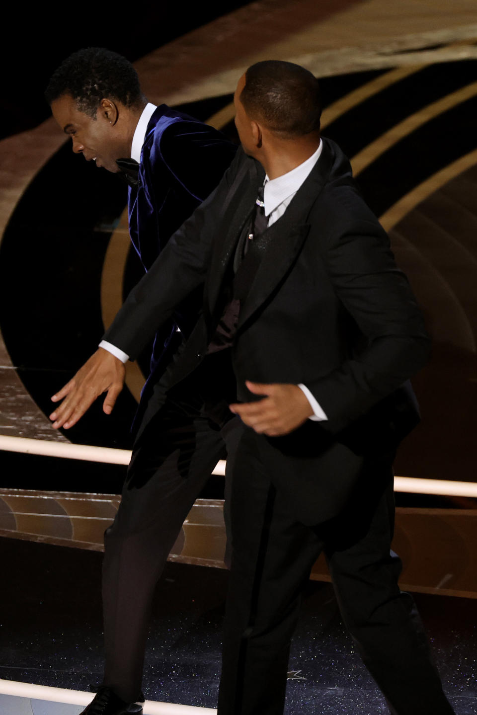 Will Smith in a black tuxedo appears to be hitting Chris Rock, who is also in a tuxedo, on stage at an awards show