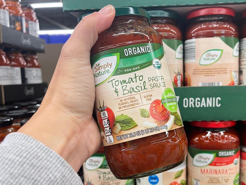 A hand holding a jar of Simply Nature tomato and basil pasta sauce in front of a display of other jars.