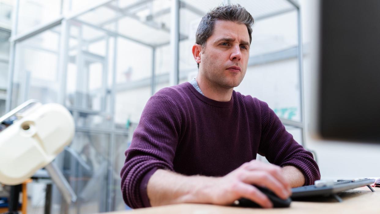 A man wearing a purple sweater in front of a desktop computer with his hand on a computer mouse, looking intensely at the screen.