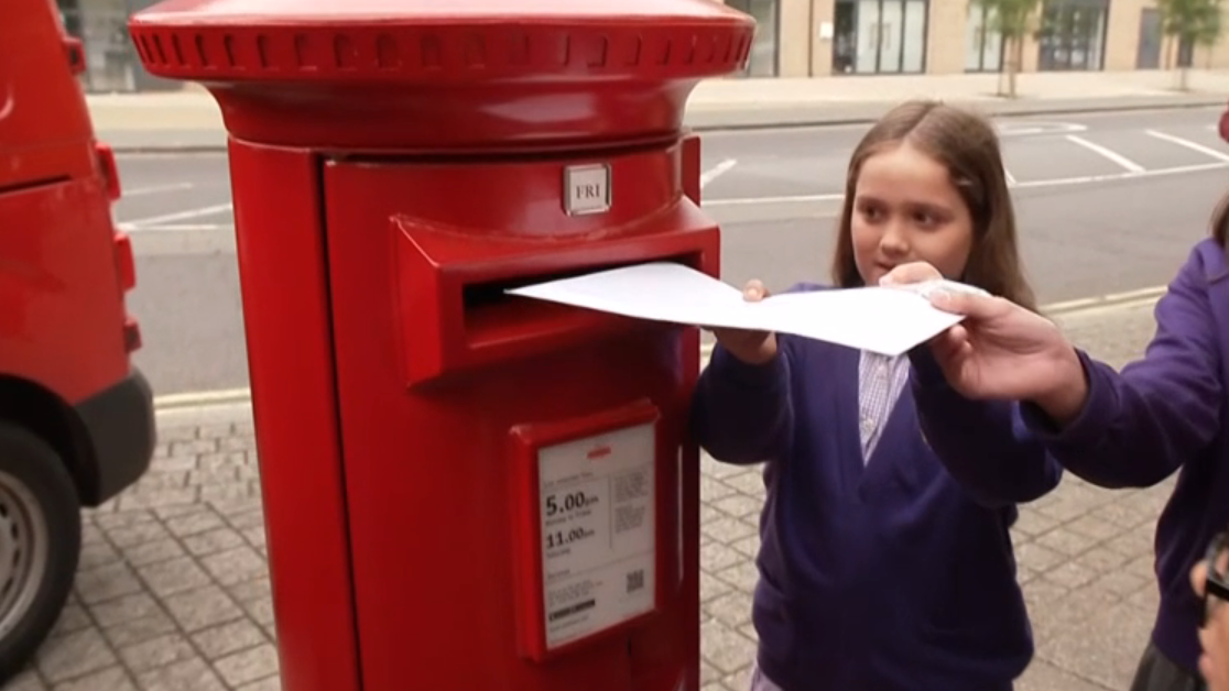A young girl helps an adult post a white envelope into a red post box on a street