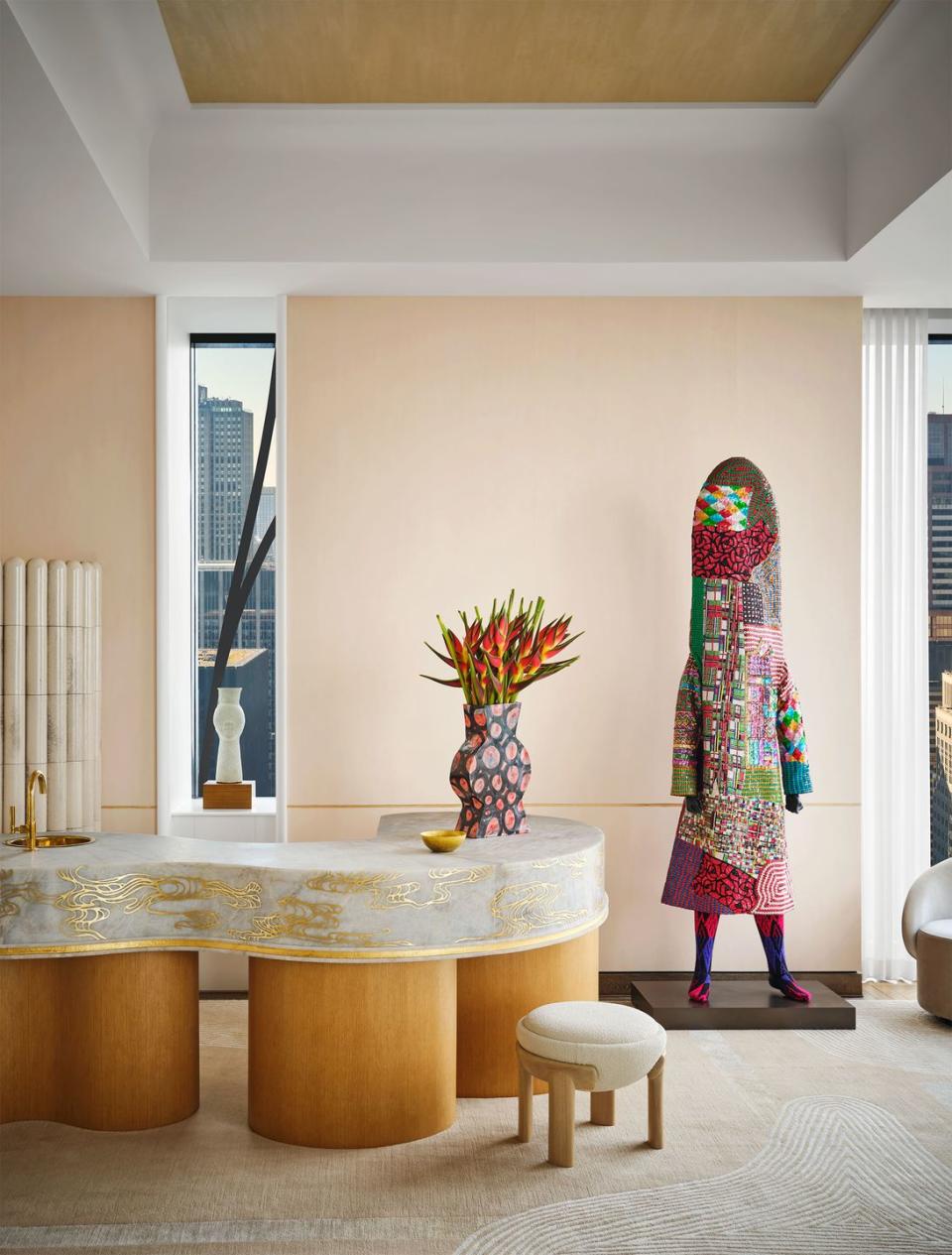 a multicolored human size sculpture stands next to a curved wet bar with a quartzite top and swirly gold pattern, papier mache vase with flowers, narrow window with an artful vase on sill