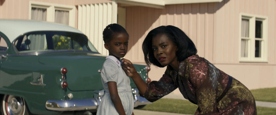 Melody Hurd stands in front of a house and car next to Deborah Ayorinde, who bends over to touch her