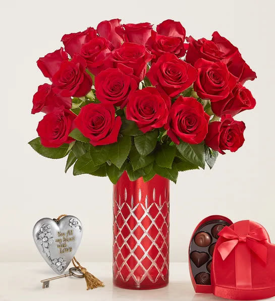 24 red roses in red vase with silver heart decor nearby along with a heart shaped box of chocolates. 