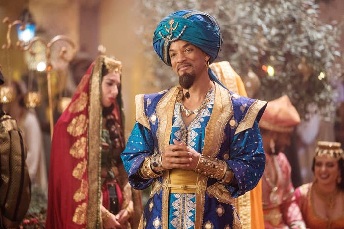 Will Smith dressed as Genie from Aladdin, wearing ornate blue and gold attire with a turban, stands with a content expression amidst a festive setting