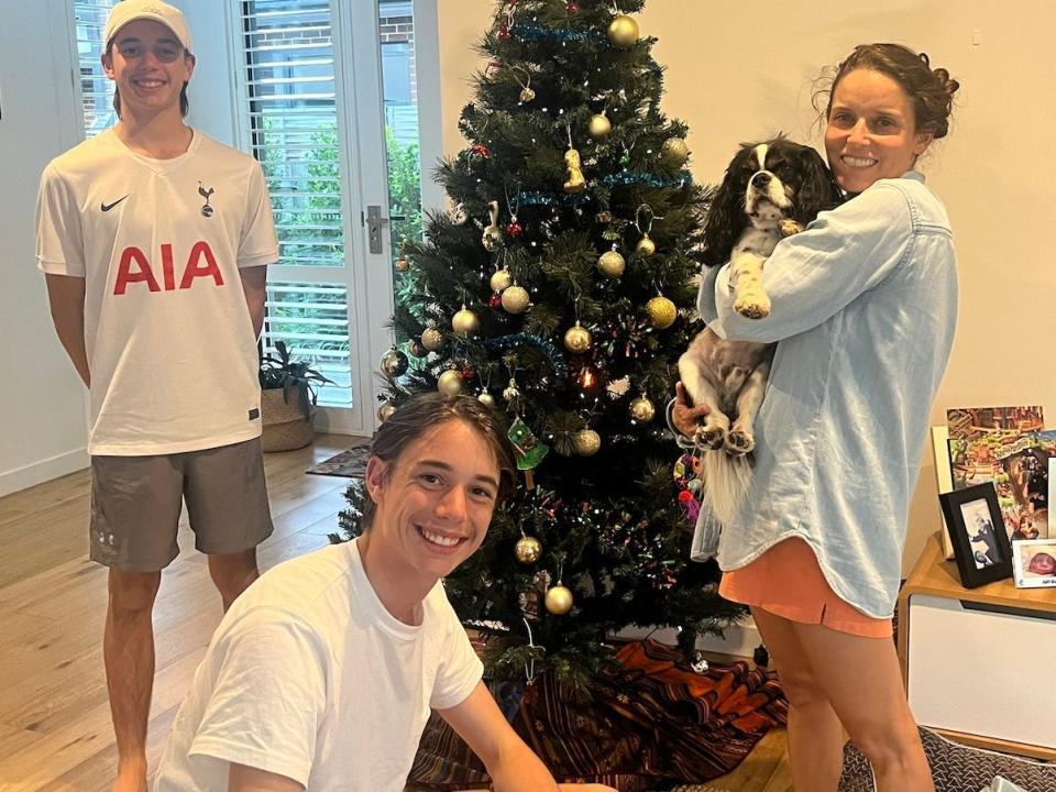 Photo of Ash Jurberg's sons, Charlie and Thomas, and his wife in front of a Christmas tree. One of the twins squats in front of the Christmas tree and is angled to the viewer's right, but smiles at the camera, wearing a white t-shirt and black shorts. The other twin stands behind his brother with his hands behind his back He wears a white baseball cap, white athletic shirt and grey shorts. Ash's wife Cecelia stands to the right of the Christmas tree and holds a black and white King Charles Spaniel dog. She wears a denim shirt, salmon pink shorts, and smiles widely. All three have dark brown hair and dark eyes.