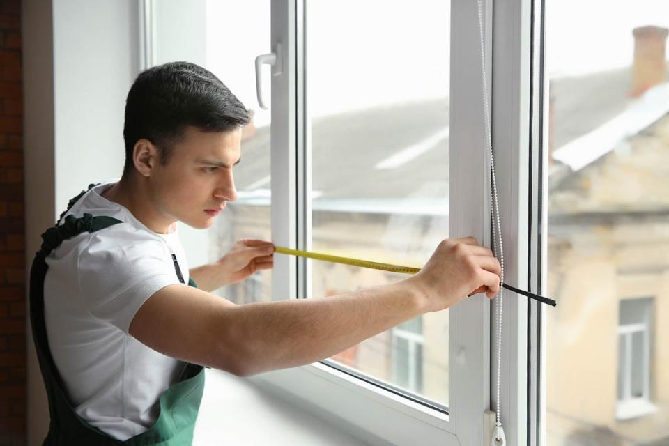 A man in green overalls measures the width of a window from the inside.