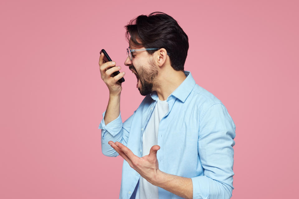 Image of aggressive young man in blue shirt screaming at smartphone while having mobile conversation isolated over pink background