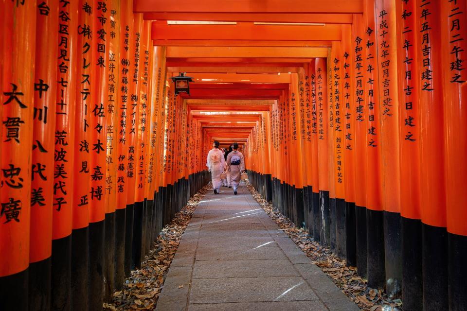 Women in traditional kimonos walk through red gates with Japanese writing written on them