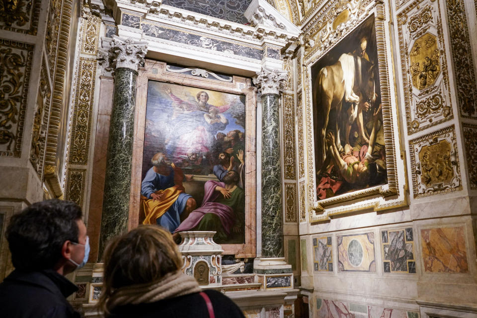 Visitors admire the "Conversion (of St. Paul) on the Way to Damascus", 1601, right, by Renaissance master Michelangelo Merisi, known as Caravaggio of 1601, flanking "The Assumption of the Virgin", 1601, by Annibale Carracci, inside the Cerasi Chapel of Santa Maria del Popolo church, Rome, Friday, Dec. 11, 2020. Like elsewhere in Europe, museums and art galleries in Italy were closed this fall to contain the spread of COVID-19, meaning art lovers must rely on virtual tours to catch a glimpse of the treasures held by famous institutions such as the Uffizi in Florence and the Vatican Museums in Rome. However, some exquisite gems of Italy's cultural heritage remain on display in real life inside the country's churches, some of which have collections of renaissance art and iconography that would be the envy of any museum. (AP Photo/Andrew Medichini)