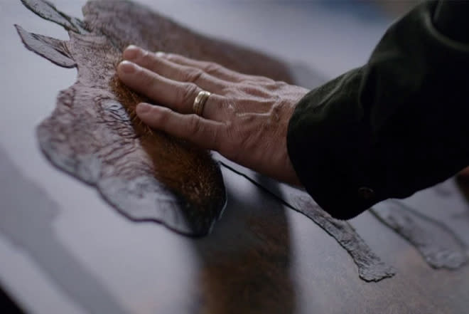 The fingertips of a hand wearing a gold wedding band gently rest on a tactile image of a rhino, printed on a flat white background. The shape of the animal is in relief and the differing textures of its skin are apparent.