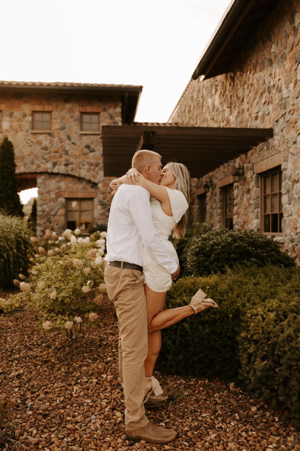 A woman in a white dress and a man in a white shirt and khakis kiss as he picks her up in front of a stone building.