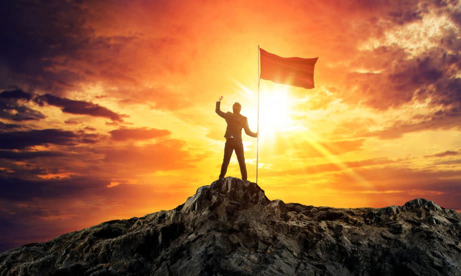 Geschäftsmann auf dem Gipfel eines Berges, in der Hand eine Fahne, im Hintergrund die untergehende Sonne