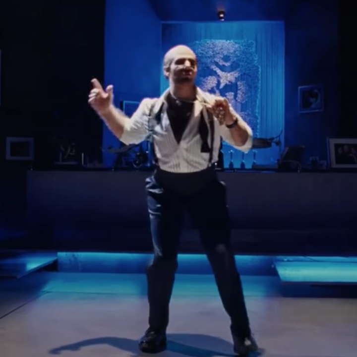 Tom Cruise in a bald cap and prosthetic, shirt unbuttoned and dancing