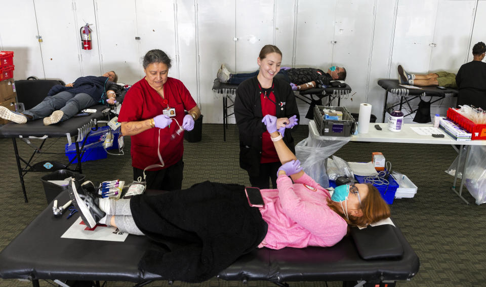 Registered Dietitian, Sheri Weitz, from Westchester, Calif., wears a face mask and gloves, as she donates blood with her husband, Gary "Robin" Wietz, seen in background, center, at the American Red Cross office in Santa Monica, Calif., Thursday, March 26, 2020. The new coronavirus causes mild or moderate symptoms for most people, but for some, especially older adults and people with existing health problems, it can cause more severe illness or death. (AP Photo/Damian Dovarganes)