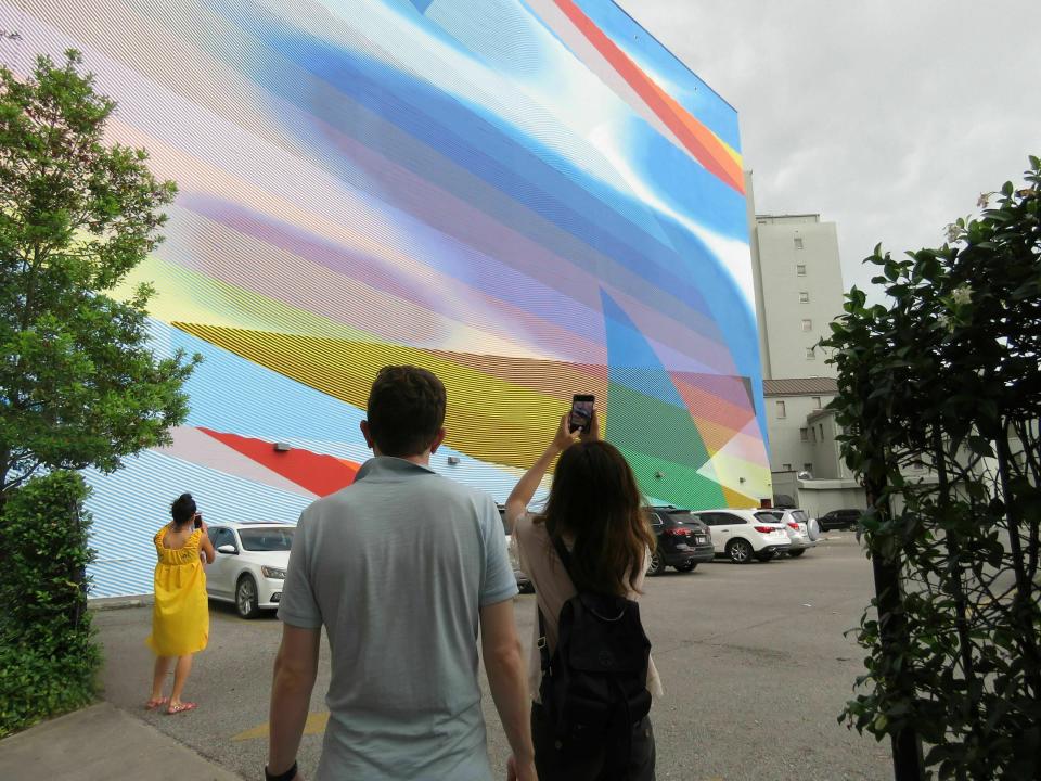 In this Thursday, May 30, 2019 photo, Cathy Ho of New York, visiting New Orleans with Matthew Lloyd of New York, takes a photo of the mural by Momo covering a side wall of the Ogden Museum of Southern Art. The mural is one of five created as part of an Arts Council of New Orleans project called "Unframed." The council's executive director, Heidi Schmalbach, says the group wants New Orleans to be known for its contemporary art as much as for its music, food and culture. (AP Photo/Janet McConnaughey)