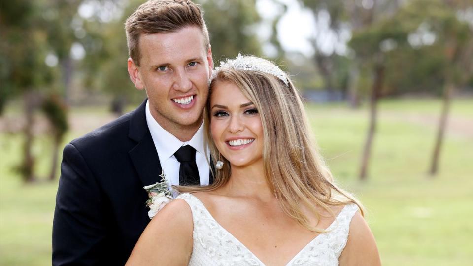 2021 MAFS couple Georgia and Liam in a garden at their show wedding. He is clean shaven in a dark suit and tie. She wears a tiara and a lace gown with V-neckline and her streaked hair free to her shoulders.