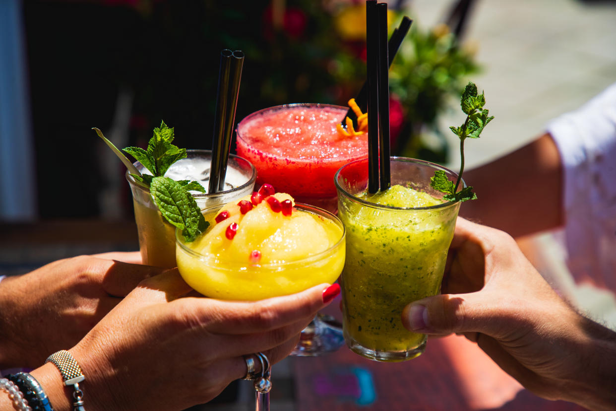 Four hands holding drinking glasses with yellow and red fruit cocktails, proposing a toast at outdoor party