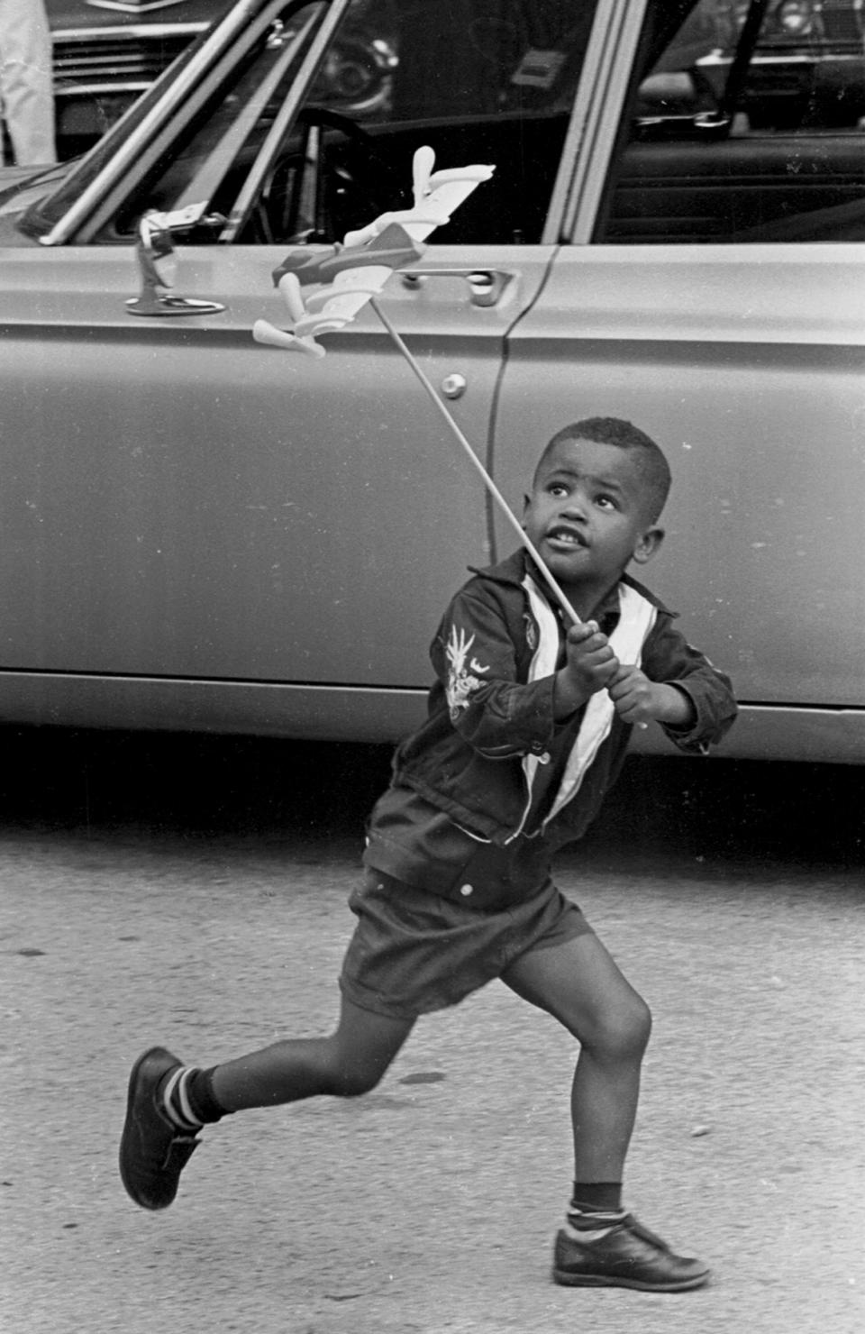 A young boy is intently focused upon his toy airplane, perched on a long stick, as he 'flies' it by running alongside a parked car, Chicago, 1965. (Photo by Robert Abbott Sengstacke/Getty Images)