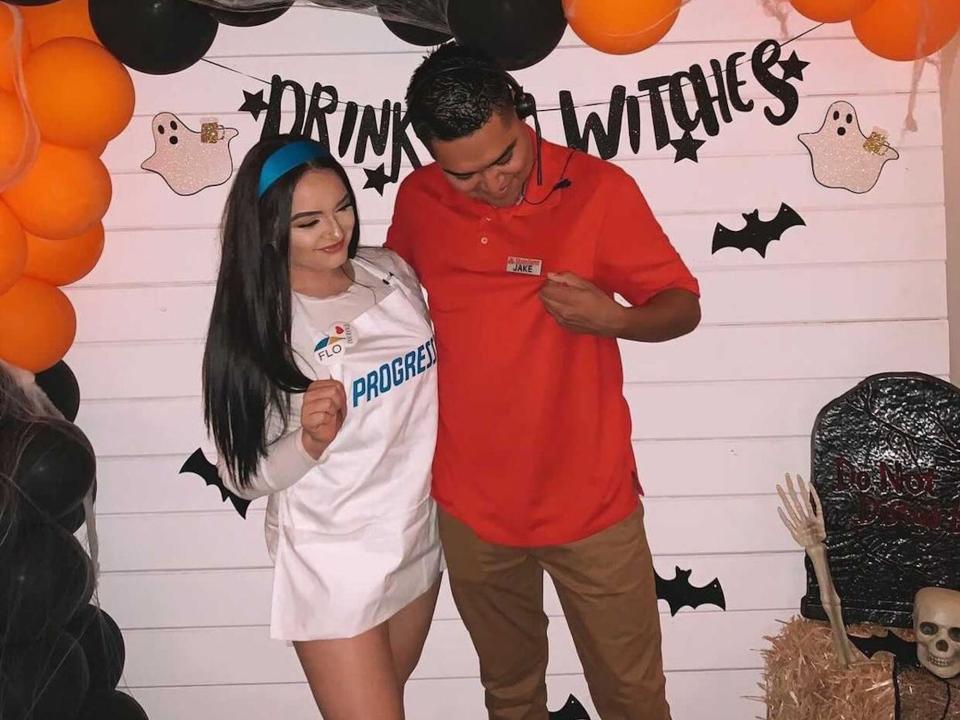 A couple dressed as Flo from Progressive and Jake from State Farm pose in front of a black and orange balloon wall.