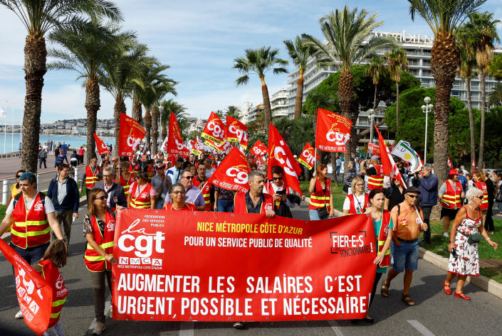 Franču arodbiedrību darbinieki staigā pa palmām ieskautu ielu, vicinot koši karogus un atbilstošu reklāmkarogu.