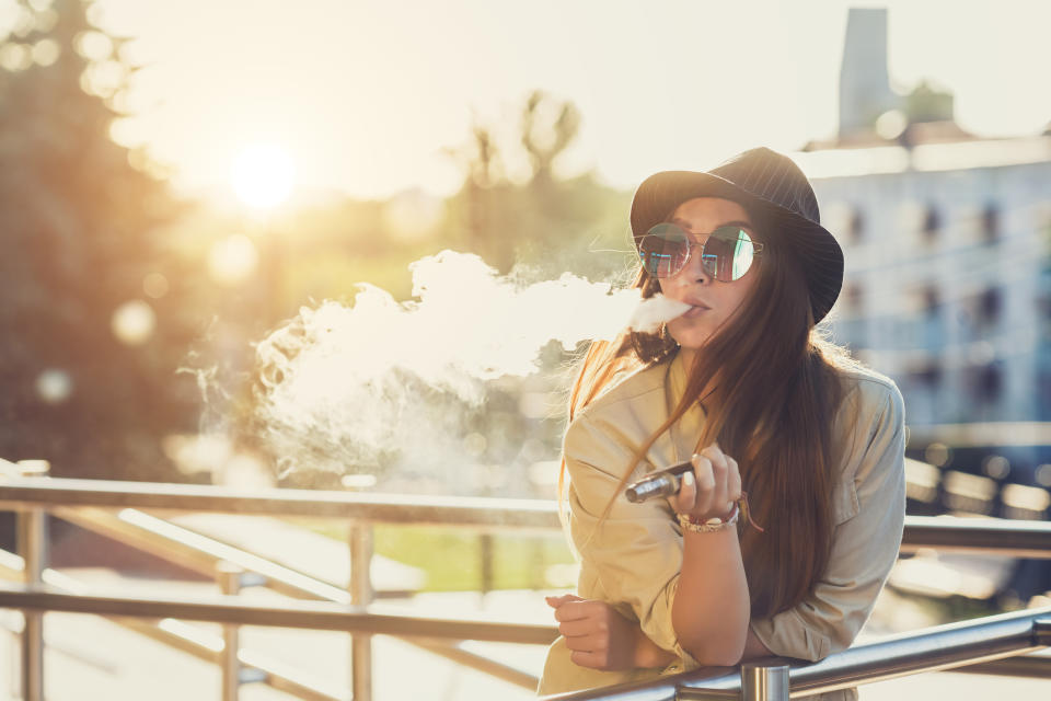 A young woman, outdoors, leaning on a railing, exhaling vapor while using an electronic cigarette.