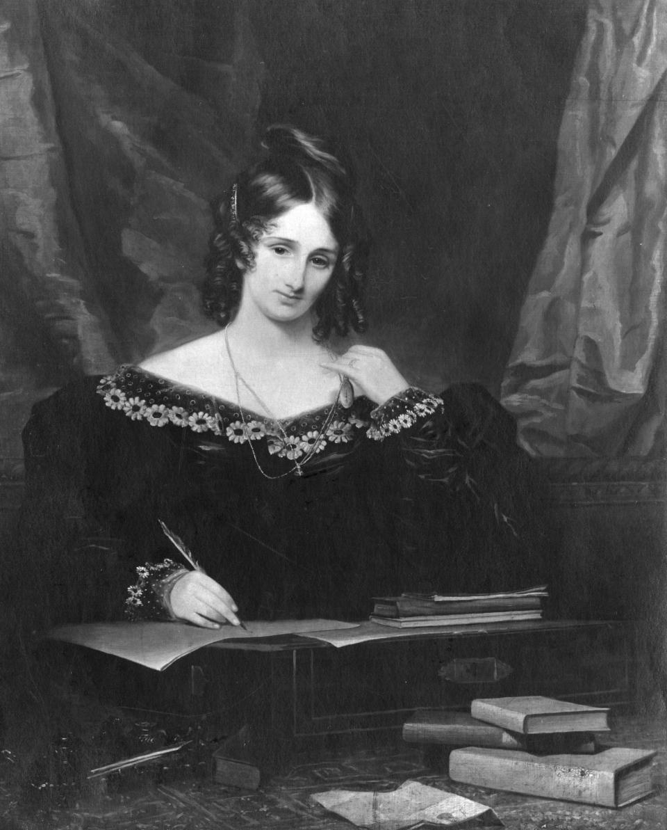 Black and white image of Mary Shelley wearing a dark dress and writing surrounded by books