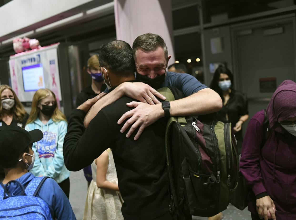 Surrounded by other people, a masked man embraces another man wearing a backpack