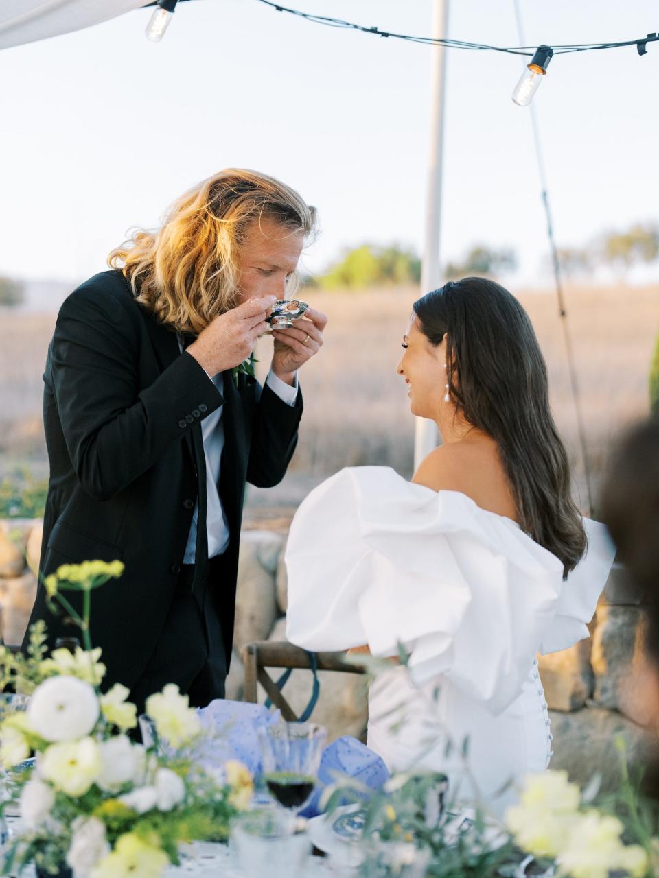 A groom drinks something as his bride watches on their wedding day.