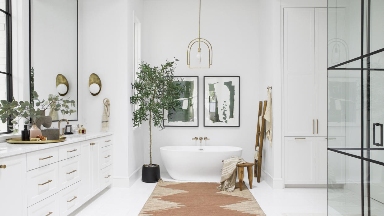  White bathroom with white countertop, white freestanding bath, potted tree, terracotta rug and brass wall sconce and ceiling pendant. 