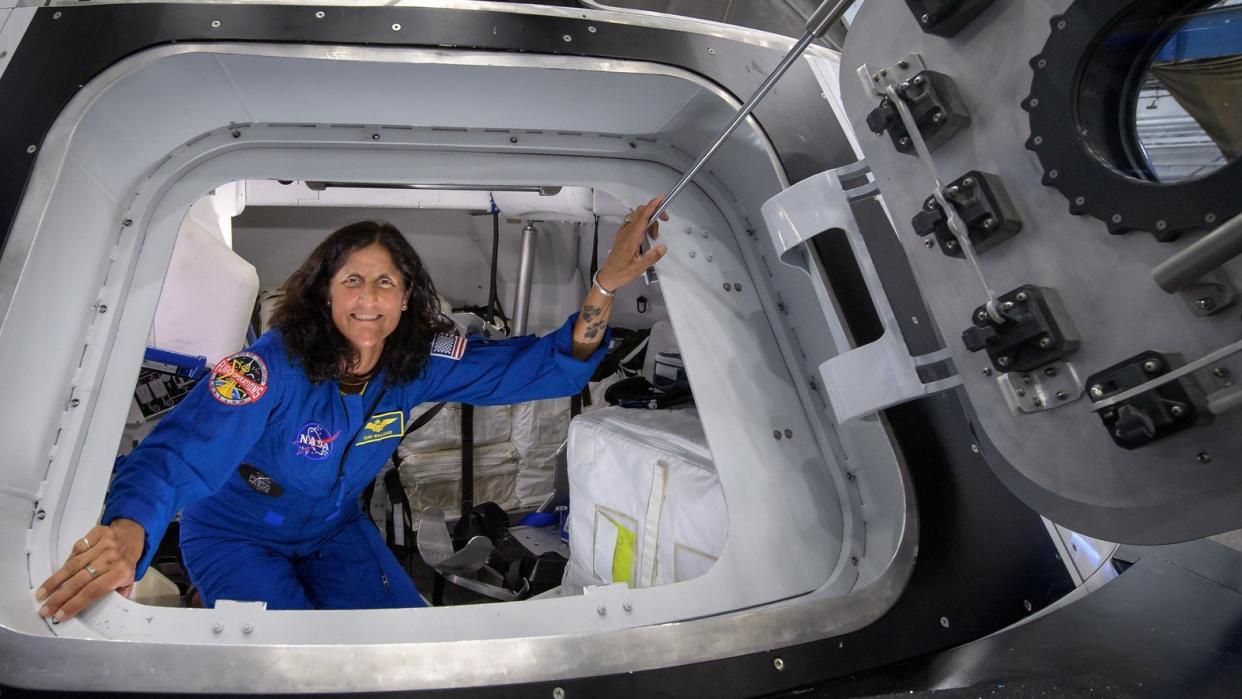  A woman astronaut in a flight suit posing in the doorway of a spacecraft simulator. the open door is at right. 