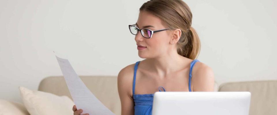 Young woman holding documents, attentively reading examining papers near laptop computer, calculating bills or checking bank statements, distance remote working and studying at home concept