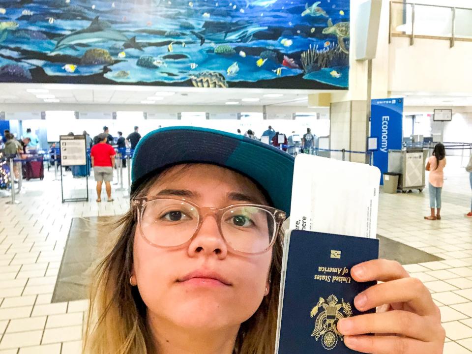 The author in a blue cap holds up her passport for a selfie inside of the airport on Guam. Behind her, there's a check-in line below a blue mural of ocean life.