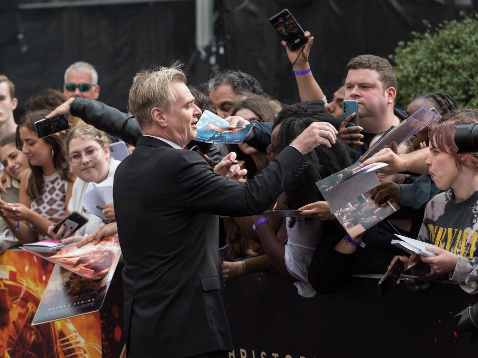 christopher nolan wearing a suit reaches into a crowd of people holding out posters at the red carpet