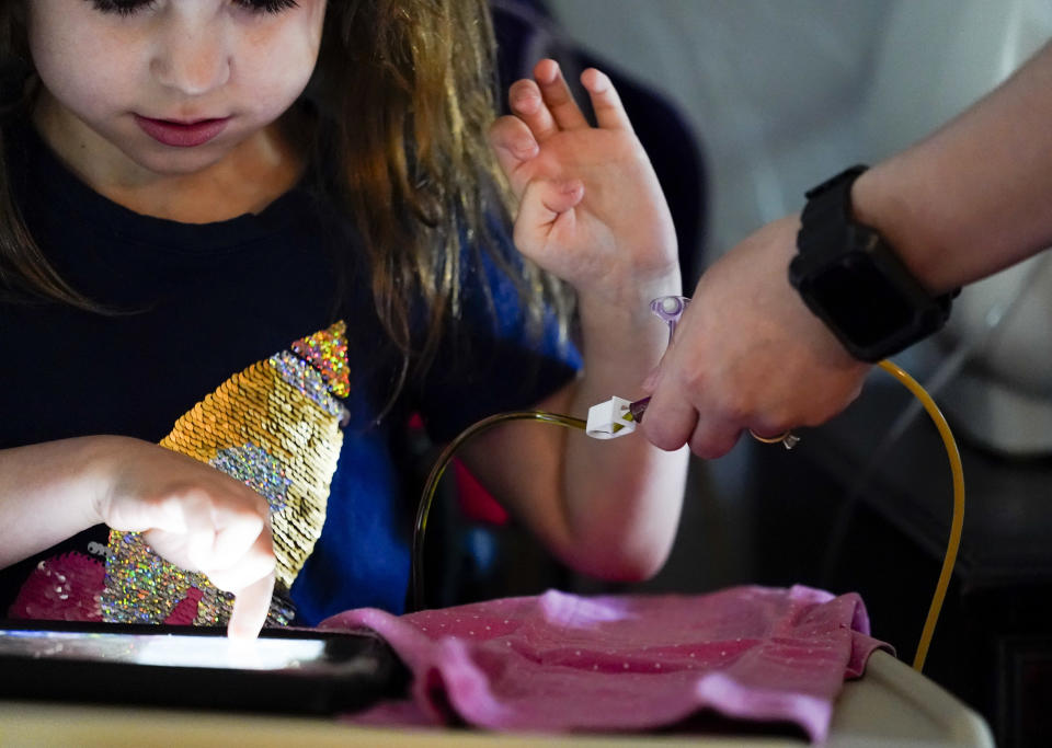 Scarlett Rasmussen, 8, watches a video on her tablet as mother, Chelsea, administers medication while they get ready for school, Wednesday, May 17, 2023, at their home in Grants Pass, Ore. Chelsea, has fought for more than a year for her daughter, Scarlett, to attend full days at school after starting with a three-day school week. She says school employees told her the district lacked the staff to tend to Scarlett’s medical and educational needs, which the district denies. Scarlett is nonverbal and uses an electronic device and online videos to communicate, but reads at her grade level. She was born with a genetic condition that causes her to have seizures and makes it hard for her to eat and digest food, requiring her to need a resident nurse at school. (AP Photo/Lindsey Wasson)