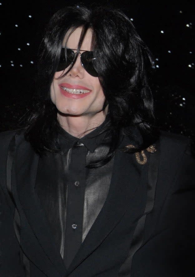 A smiling Michael in a black suit and sunglasses with shoulder-length curly hair