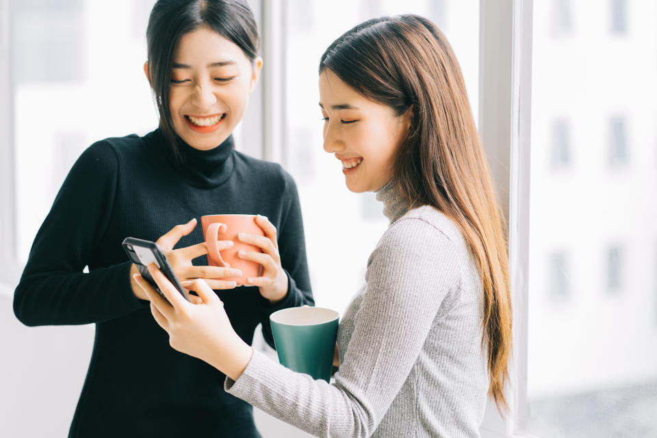Two women smile and laugh as they look at one person's phone. They are both holding mugs