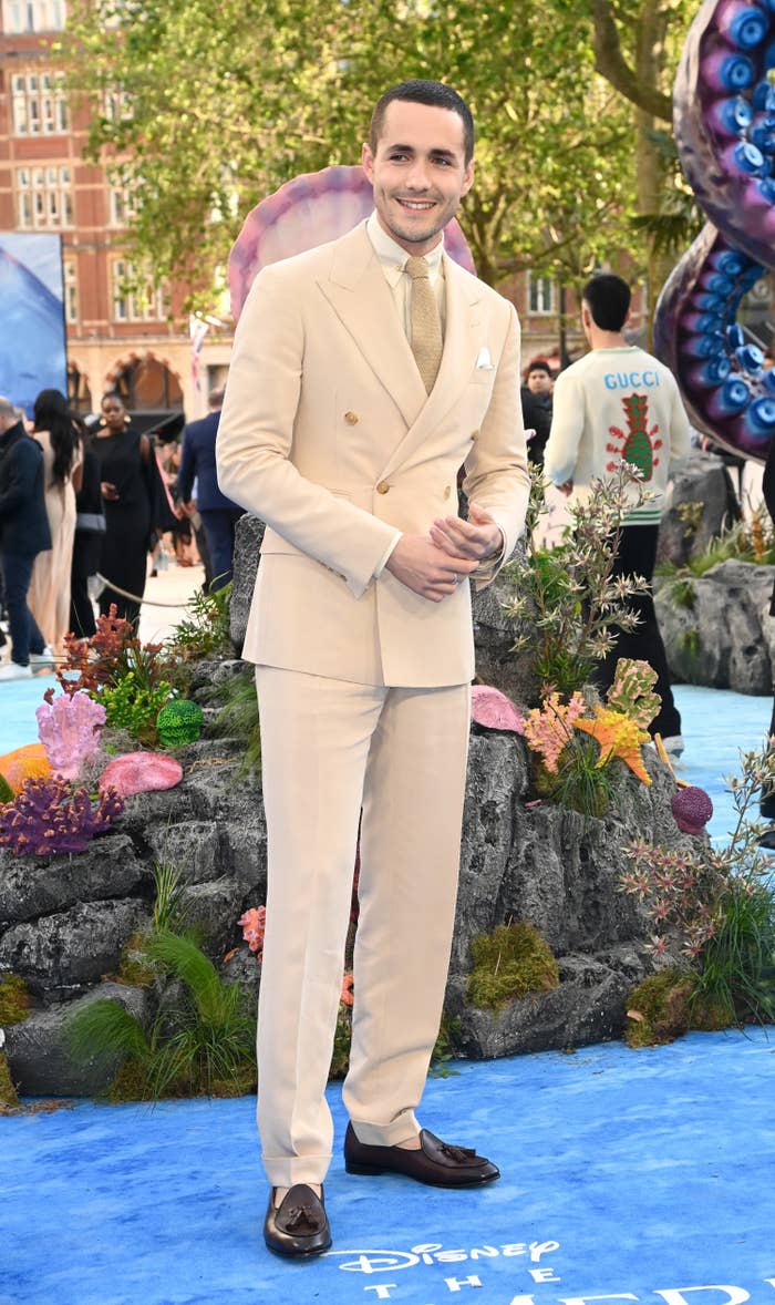 Jonah Hauer-King wears a suit in which the jacket, collared shirt, tie, and pants are all shades of the same cream color