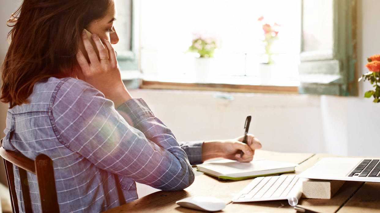 Shot of an attractive young woman writing notes while talking on a cellphone at home.