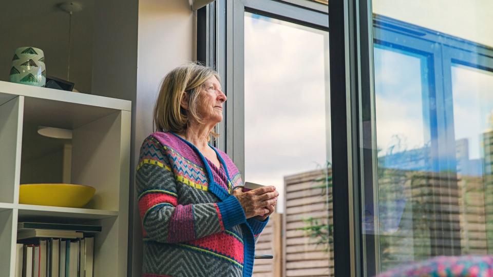 Senior woman in colourful jacket looks out window, holding cup of coffee, while in COVID-19 home quarantine. 