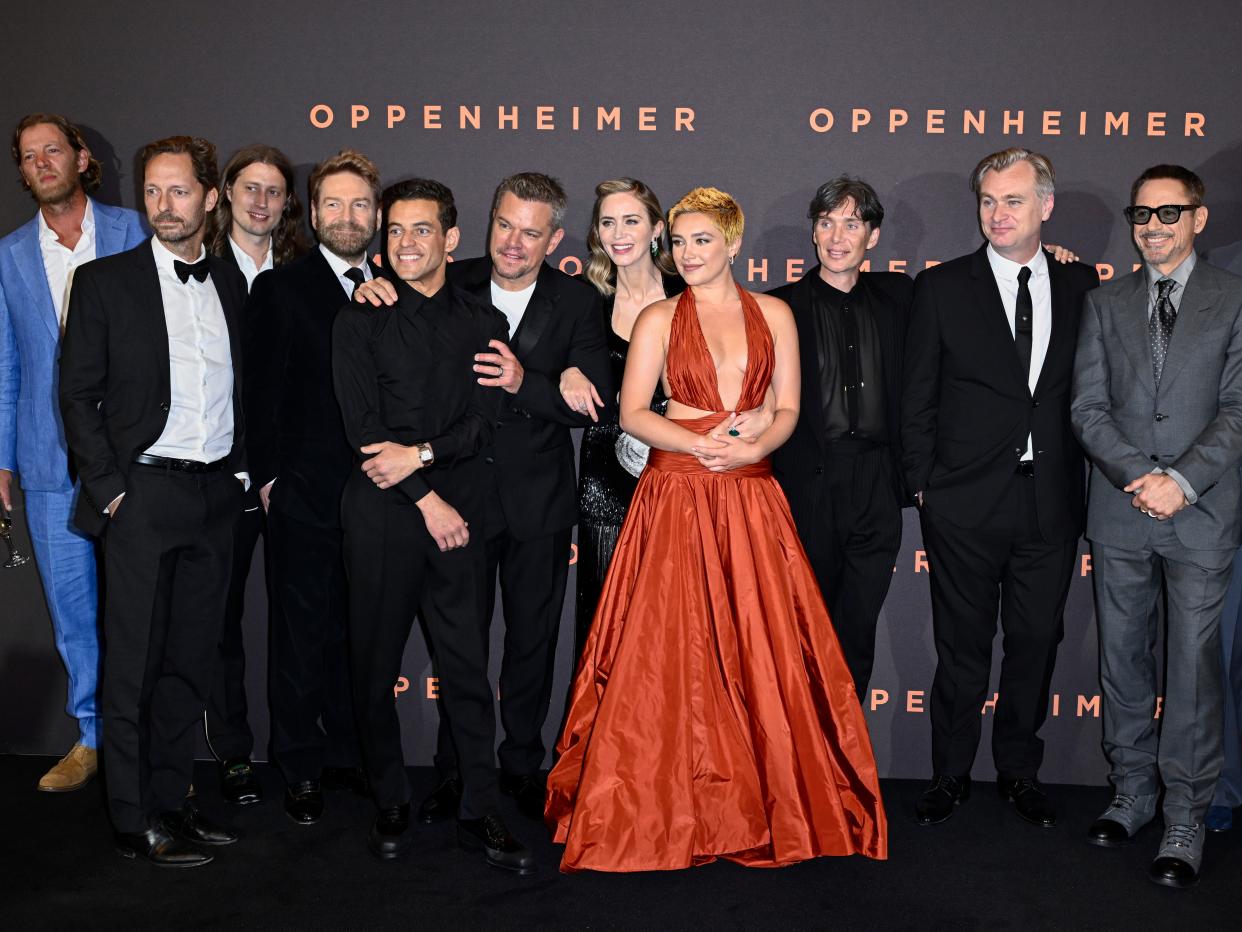 the cast of oppenheimer standing together at the film's premiere in london. florence pugh can be seen, center, with a burnt orange dress and cropped hair, standing amid cillian murphy, director christopher nolan, emily blunt, matt damon, and rami malek
