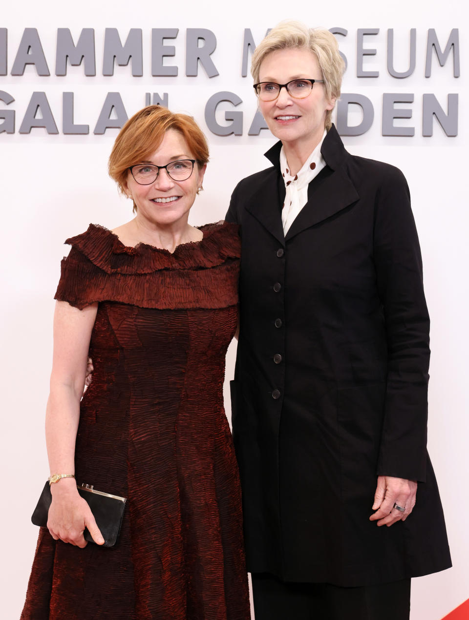 Jennifer Cheyne and Jane Lynch at the Hammer Museum Gala in the Garden, posing together. Jane Wagner is wearing a textured, knee-length dress, and Jane Lynch is in a black pantsuit with a white shirt